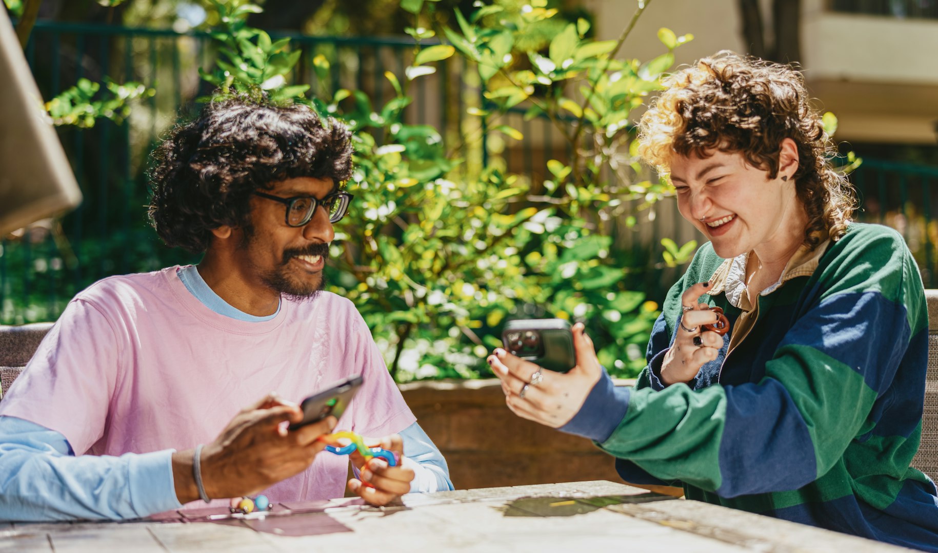 Two people holding stim toys sit together at a table. One is smiling as they show something on their phone.